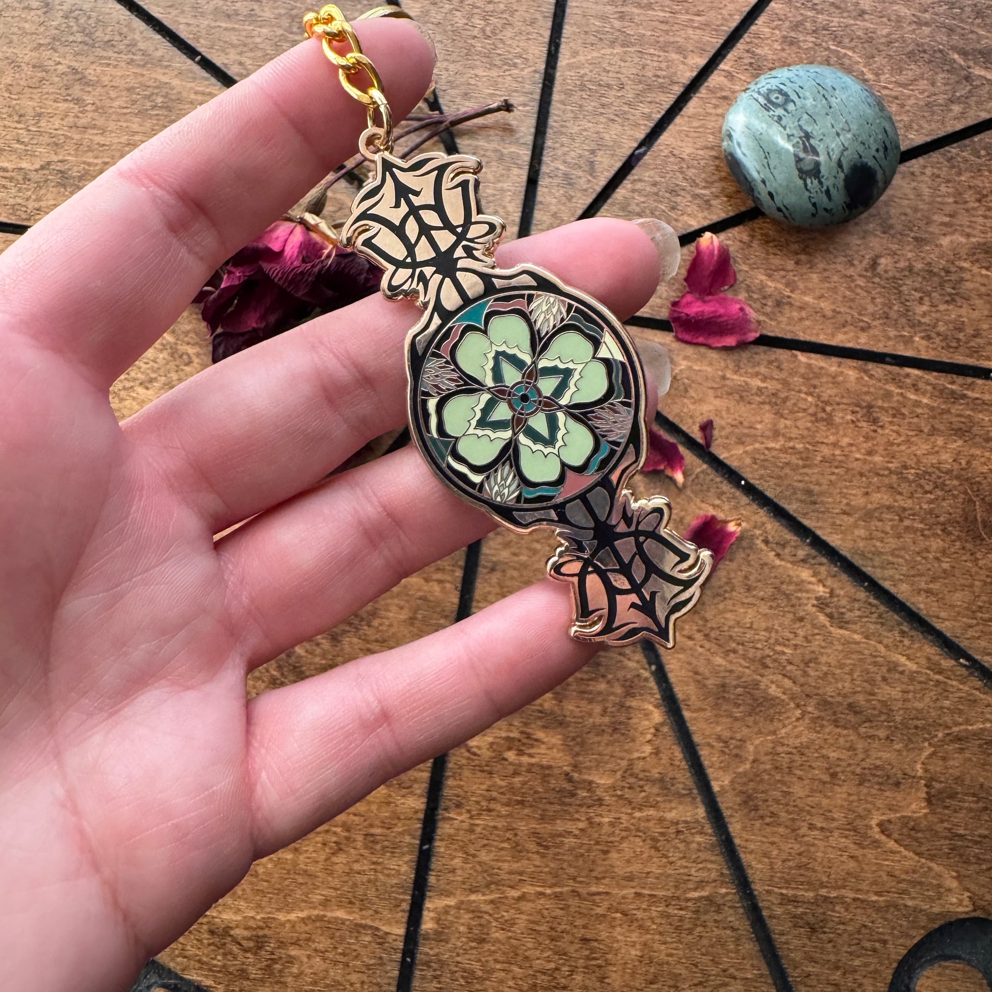 A hand holds a decorative pendant keychain with a gold-tone chain and spiderweb-shaped ends, featuring a central floral medallion with mint green, black, and blue details, over a wooden surface with dried flowers and a stone.