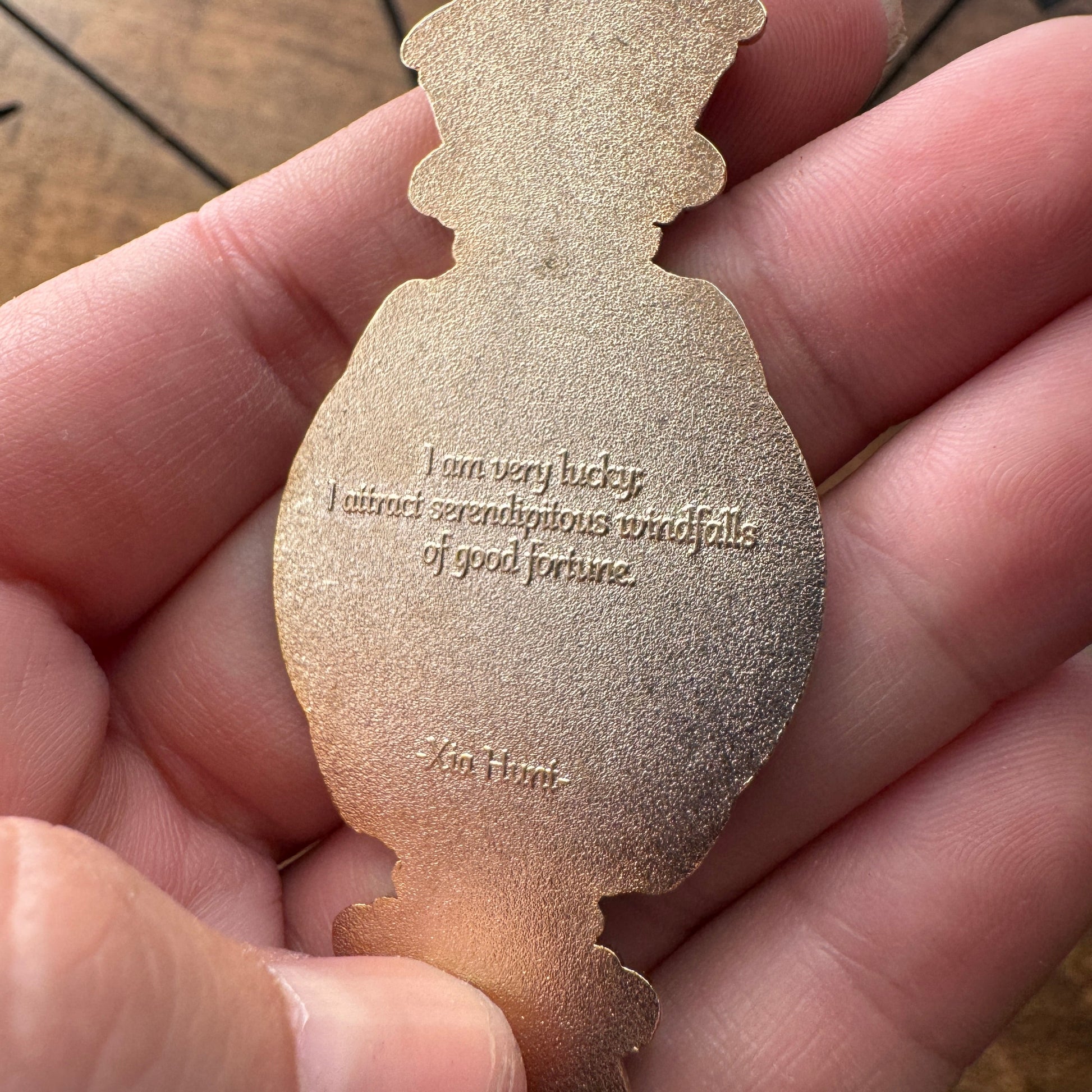 A close-up of a hand holding a metallic gold silhouette charm shaped like a perfume bottle, featuring an engraved quote by Xu Hart.