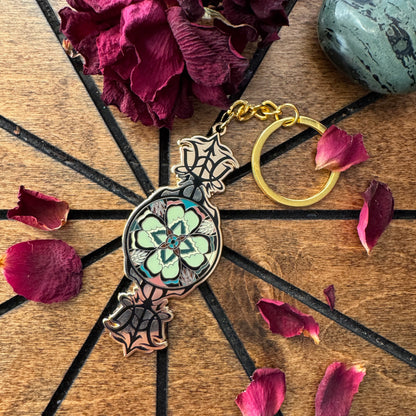 A decorative keychain with a gold-tone key ring and a central floral pendant featuring a green and black mandala design, styled on a wooden surface with dried rose petals and a stone.
