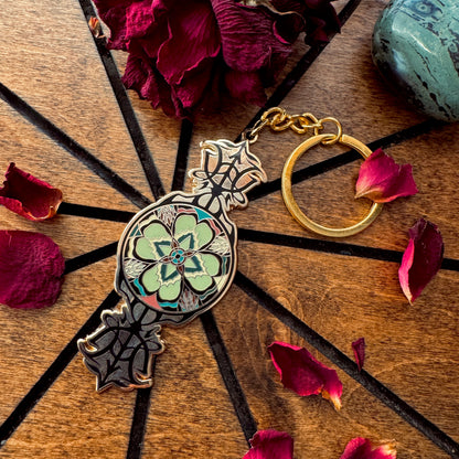 A decorative metal keychain with a floral pendant and gold key ring, styled on a wooden surface with dried rose petals and a green stone.