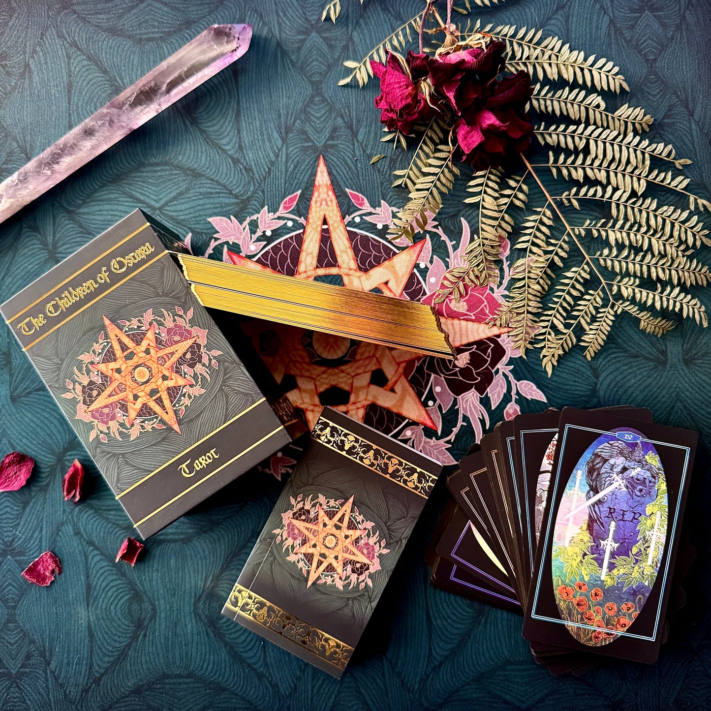 A styled flat-lay of a tarot deck set on a dark teal patterned fabric. The set includes a box labeled 'The Children of Oryx Tarot', a gold star-shaped lid, two black tarot card boxes with ornate gold and pink star designs, a fanned deck of cards showing a colorful illustration, a crystal wand, dried roses, and greenery.