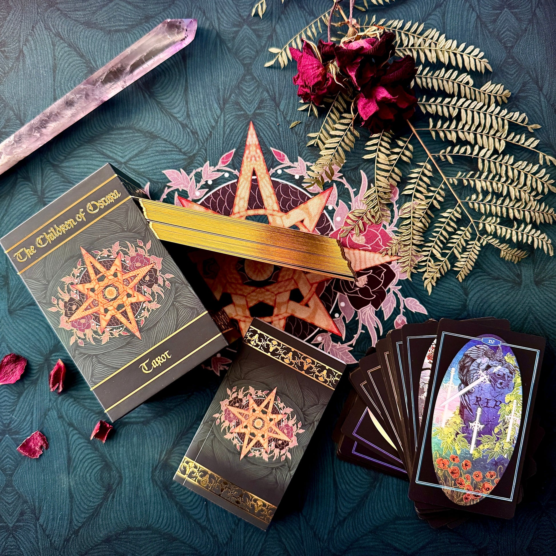 A styled flat-lay of a tarot deck set on a dark teal patterned fabric. The set includes a box labeled 'The Children of Oryx Tarot', a gold star-shaped lid, two black tarot card boxes with ornate gold and pink star designs, a fanned deck of cards showing a colorful illustration, a crystal wand, dried roses, and greenery.