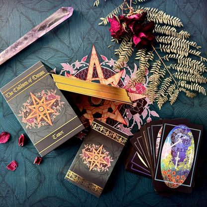 A styled flat-lay of a tarot deck set on a dark teal patterned fabric. The set includes a box labeled 'The Children of Oryx Tarot', a gold star-shaped lid, two black tarot card boxes with ornate gold and pink star designs, a fanned deck of cards showing a colorful illustration, a crystal wand, dried roses, and greenery.