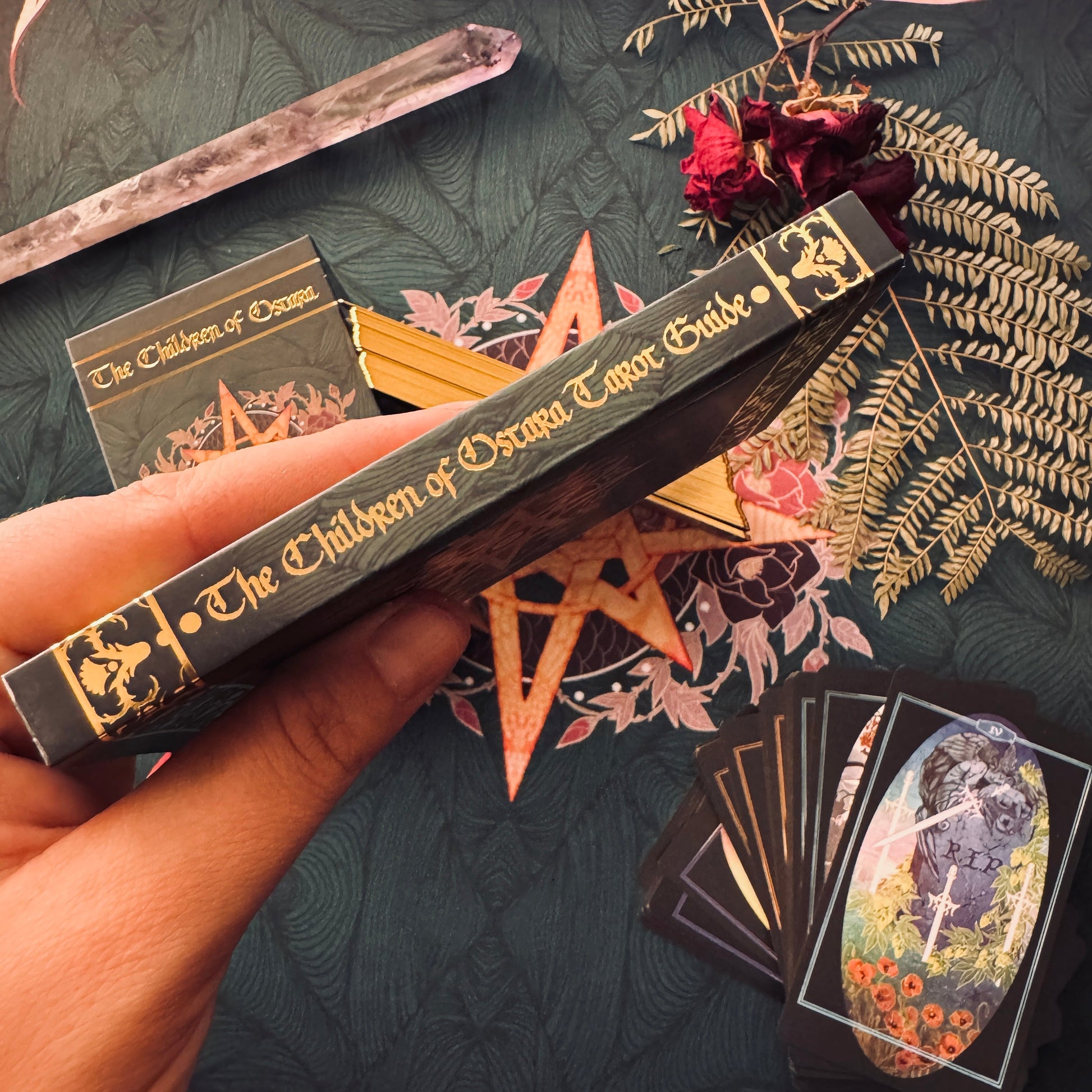 A hand holds a dark tarot deck box labeled 'The Children of Drama Editor Guide' against a dark patterned fabric background with mystical props including a crystal wand, dried red flowers, a pentagram, and tarot cards.