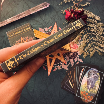A hand holds a dark tarot deck box labeled 'The Children of Drama Editor Guide' against a dark patterned fabric background with mystical props including a crystal wand, dried red flowers, a pentagram, and tarot cards.