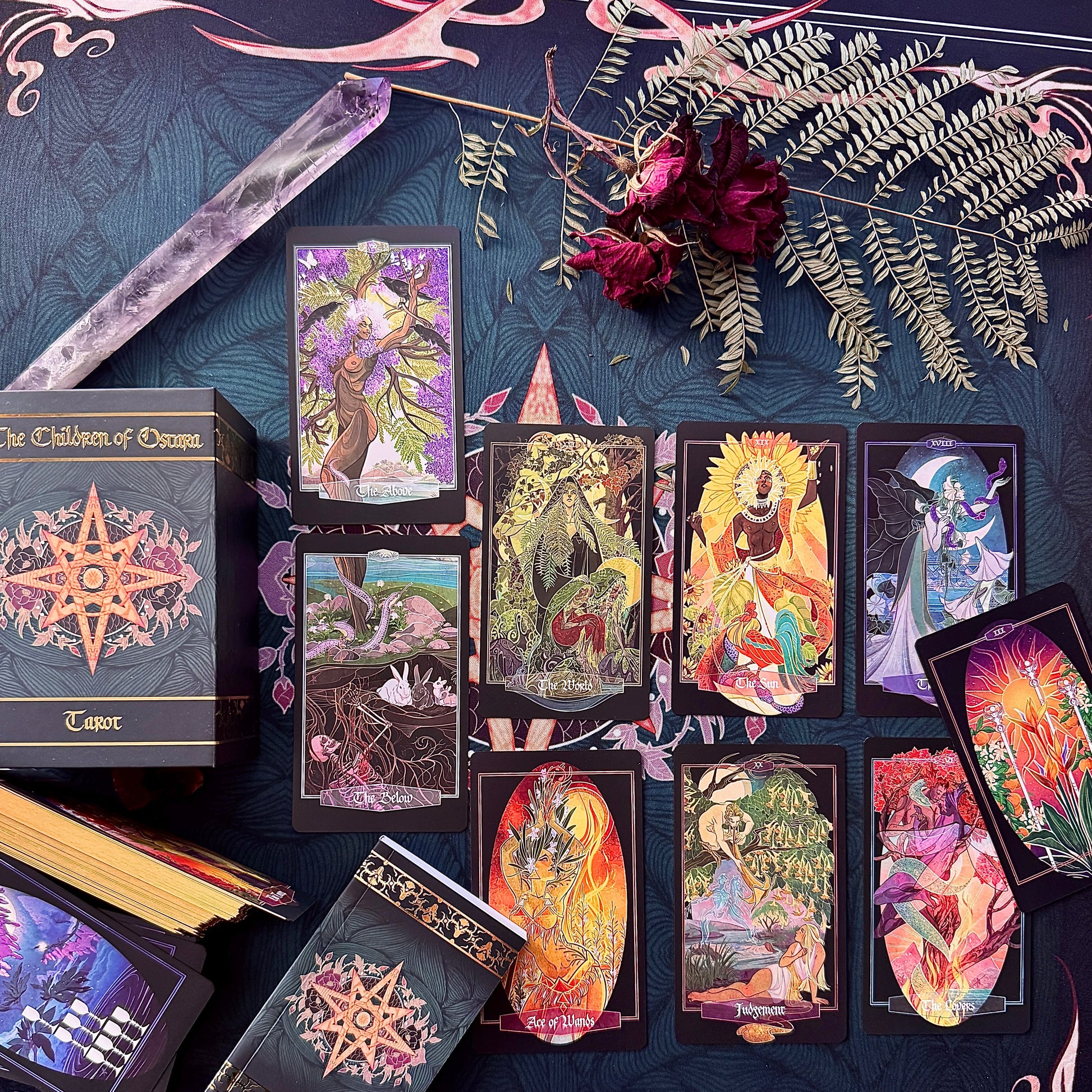 A styled flat-lay of The Children of Osiris Tarot deck, showing the main box, several cards laid out, a crystal ball, dried roses, and greenery on a dark patterned fabric background.