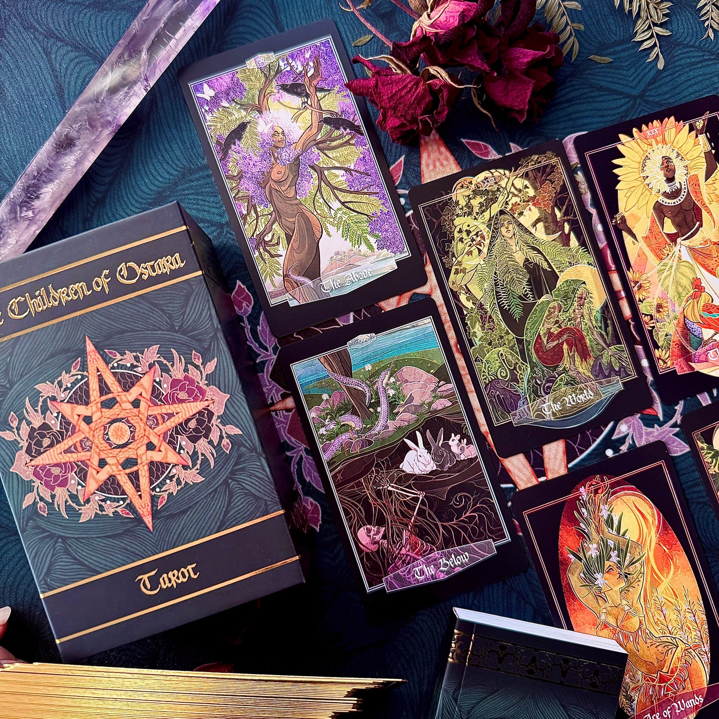 A styled flat-lay of tarot card products on a dark blue patterned fabric background, including a boxed deck labeled 'Children of Oscura Tarot' and several illustrated tarot cards with fantasy artwork.