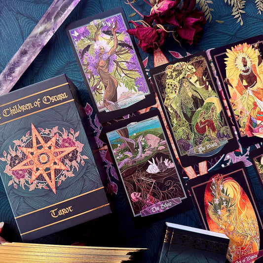 A styled flat-lay of tarot card products on a dark blue patterned fabric background, including a boxed deck labeled 'Children of Oscura Tarot' and several illustrated tarot cards with fantasy artwork.