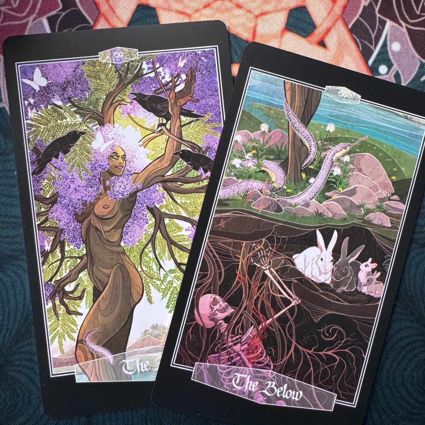 Two illustrated tarot-style cards are laid on a dark textured surface. The left card shows a tree with purple blossoms, crows, and a nude figure; the right card shows a surreal landscape with a snake, rabbits, and a skeleton in the earth.