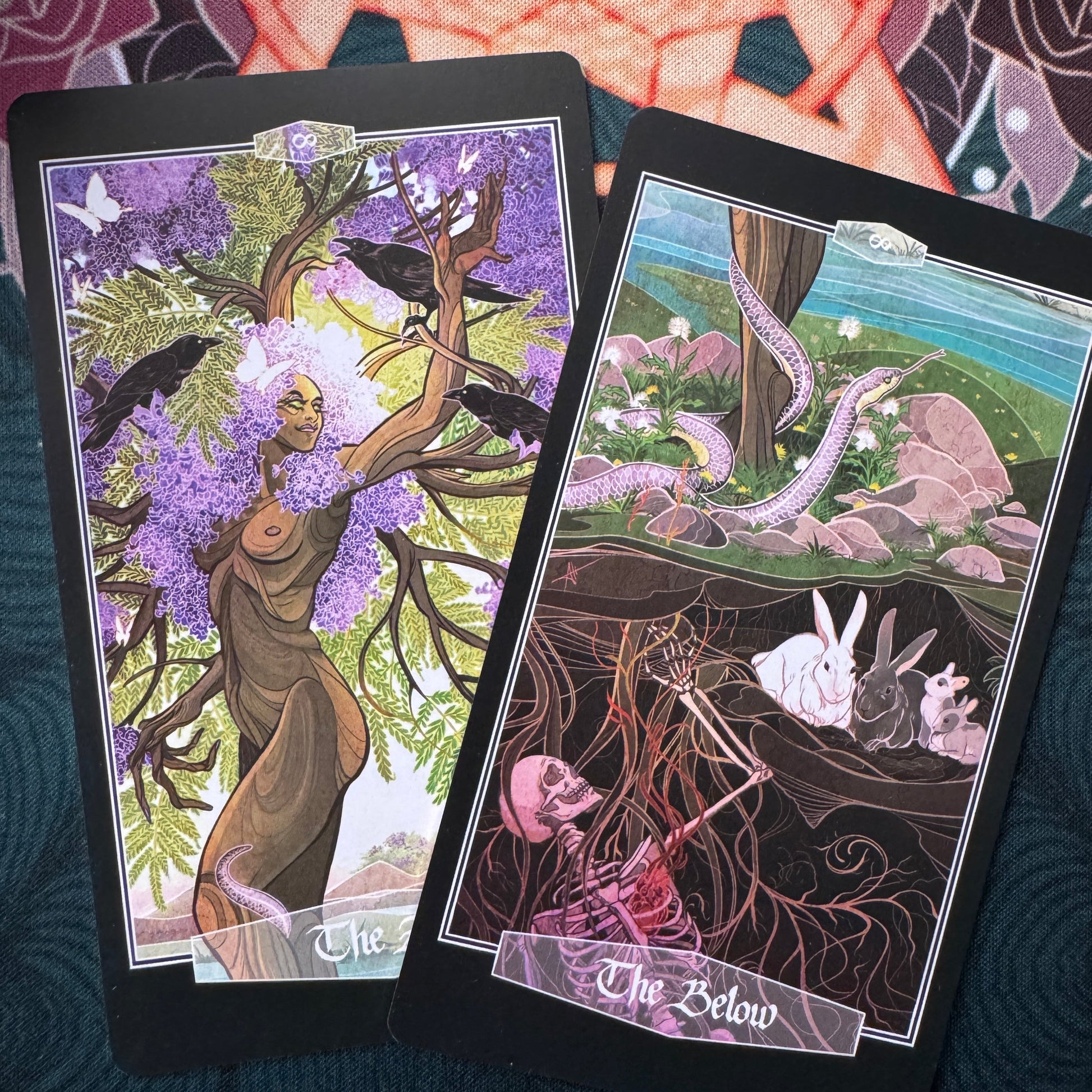 Two illustrated tarot-style cards are laid on a dark textured surface. The left card shows a tree with purple blossoms, crows, and a nude figure; the right card shows a surreal landscape with a snake, rabbits, and a skeleton in the earth.