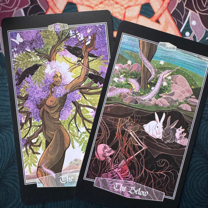 Two illustrated tarot-style cards are laid on a dark textured surface. The left card shows a tree with purple blossoms, crows, and a nude figure; the right card shows a surreal landscape with a snake, rabbits, and a skeleton in the earth.