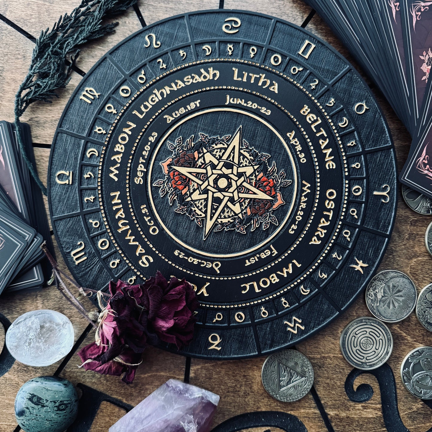 A circular black tarot card deck box or ritual board with gold zodiac and astrological symbols, surrounded by crystals, coins, dried flowers, and card packs on a wooden surface.