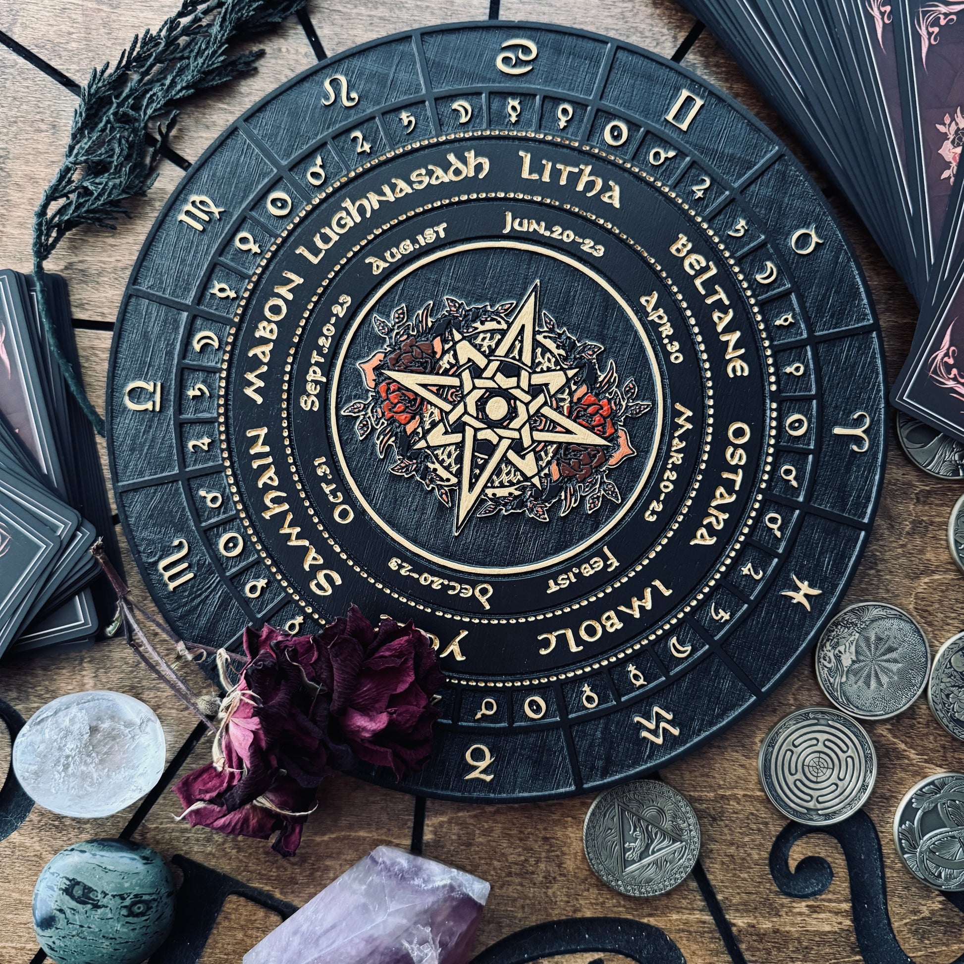 A circular black tarot card deck box or ritual board with gold zodiac and astrological symbols, surrounded by crystals, coins, dried flowers, and card packs on a wooden surface.