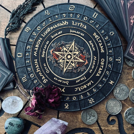A circular black tarot card deck box or ritual board with gold zodiac and astrological symbols, surrounded by crystals, coins, dried flowers, and card packs on a wooden surface.