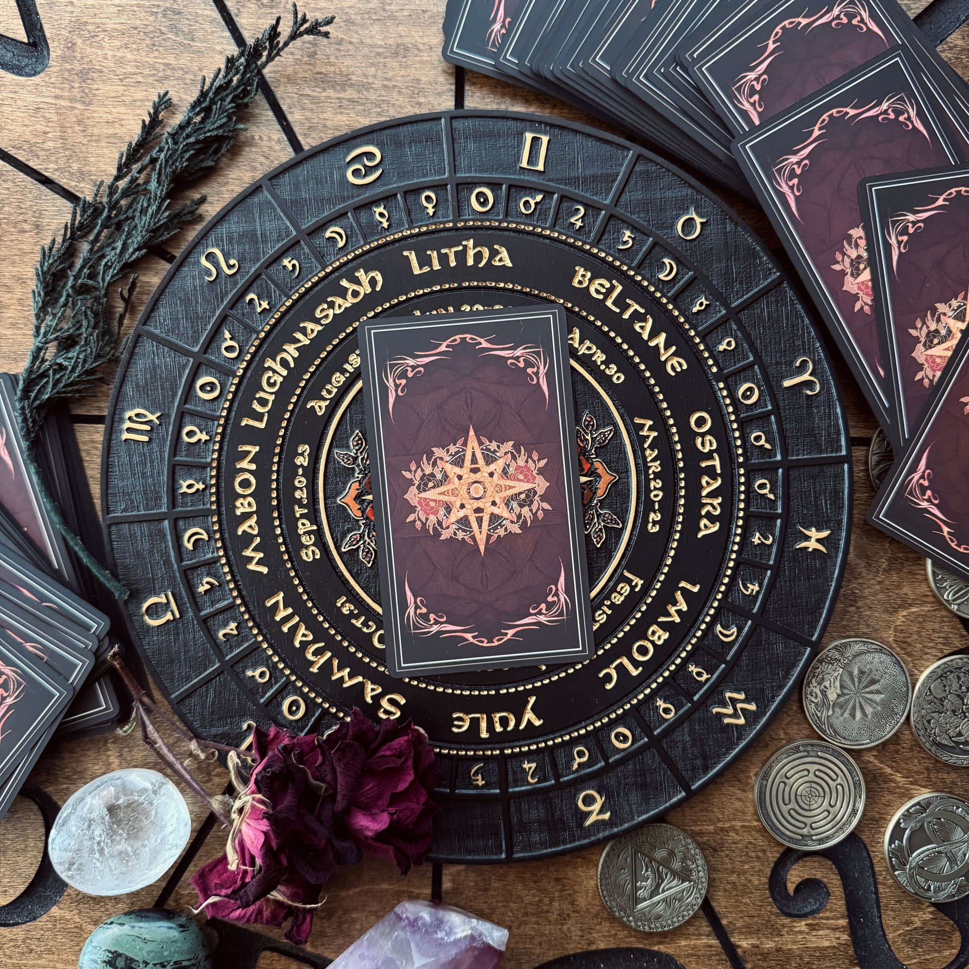 A circular black and gold Lichthaus Wiccan altar card deck set displayed on a rustic wooden surface, surrounded by crystals, coins, dried flowers, and a sprig of sage.