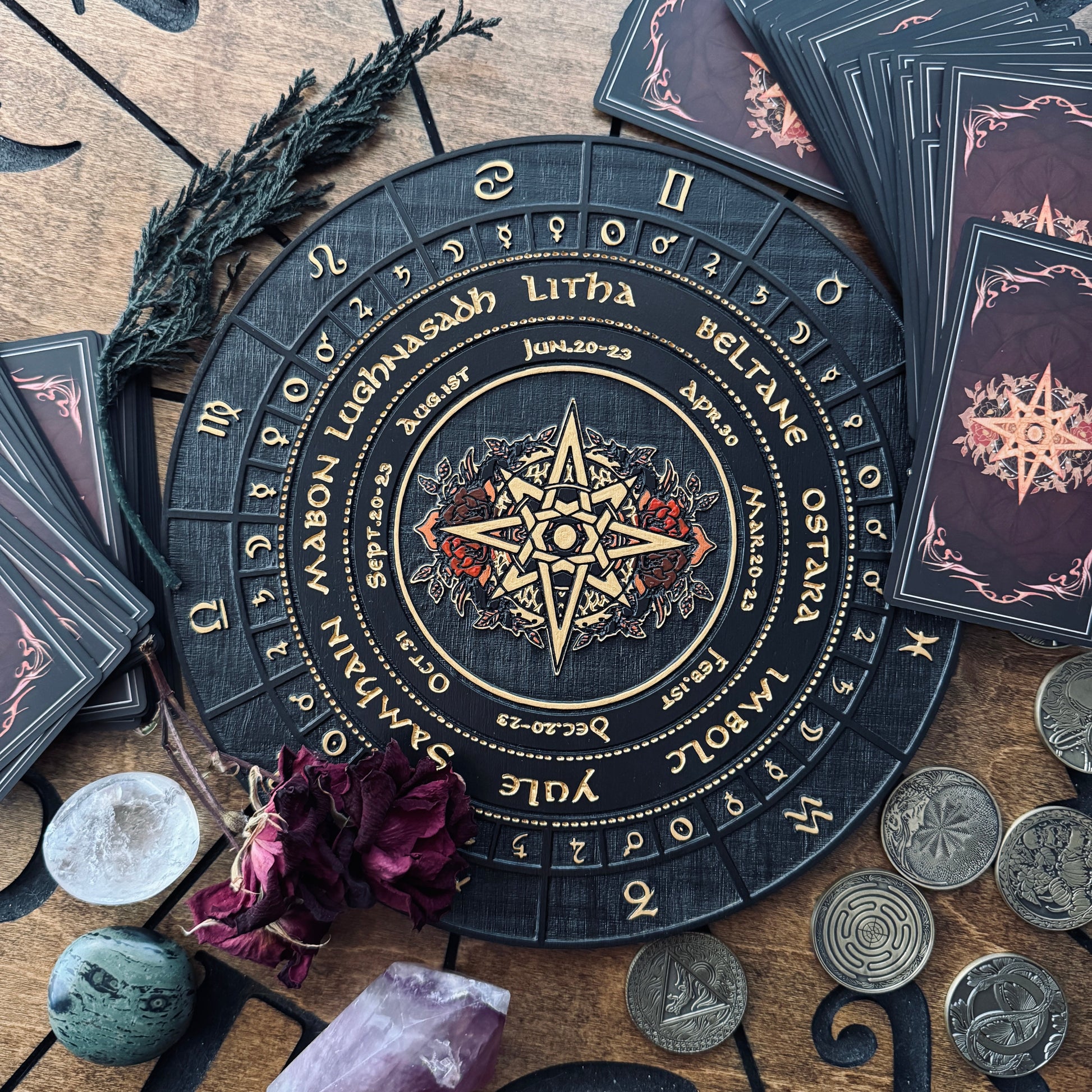 A circular tarot card wheel with zodiac signs and astrological text, surrounded by tarot cards, crystals, coins, and dried flowers on a wooden surface.
