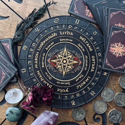 A circular tarot card wheel with zodiac signs and astrological text, surrounded by tarot cards, crystals, coins, and dried flowers on a wooden surface.