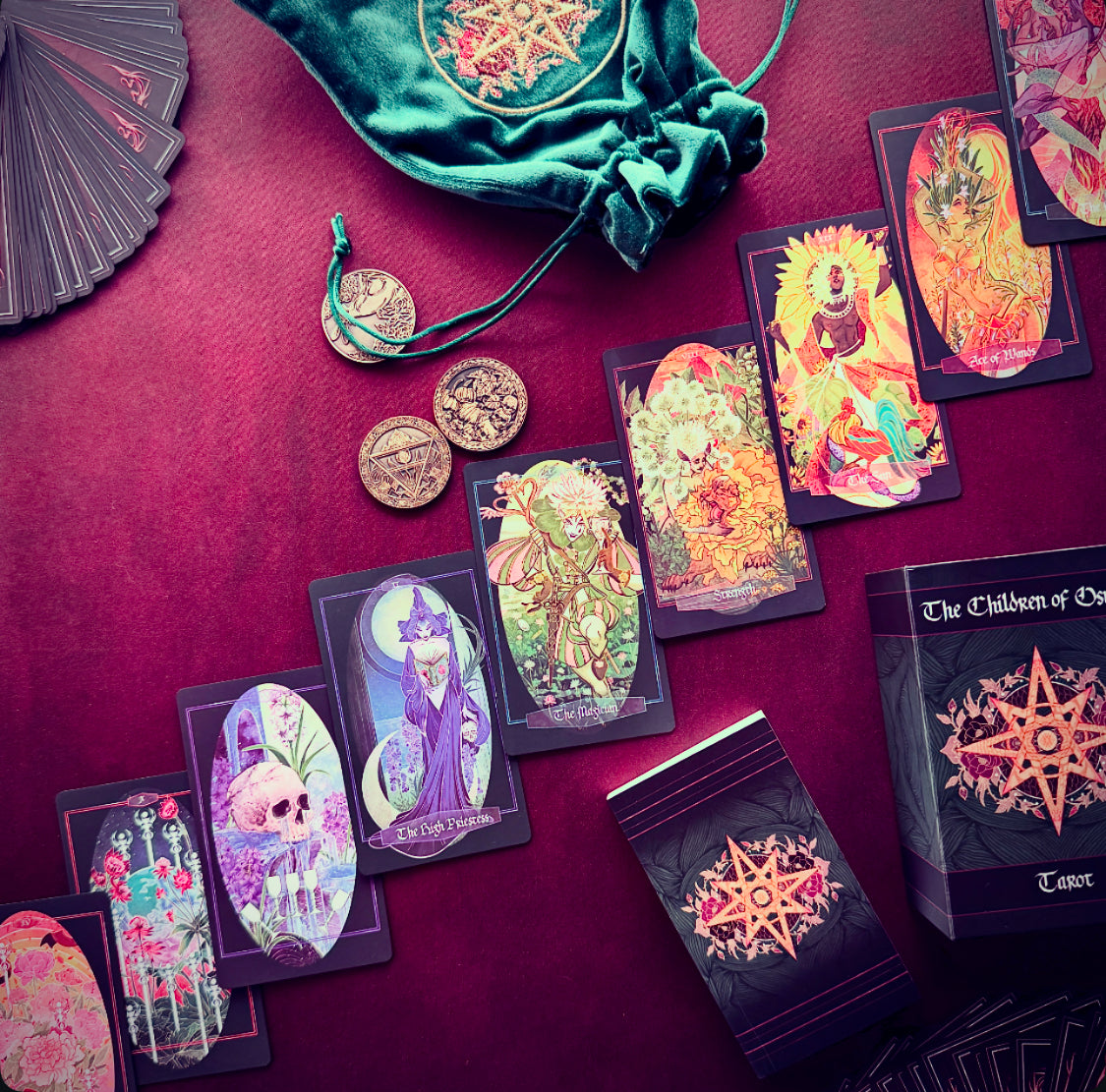 A styled flat-lay of a tarot deck set on a deep burgundy fabric background, featuring illustrated tarot cards, a green velvet drawstring bag, several gold-toned coins, and the tarot box. The cards display colorful fantasy and gothic illustrations, and the box is labeled 'The Children of Osiris Tarot'.