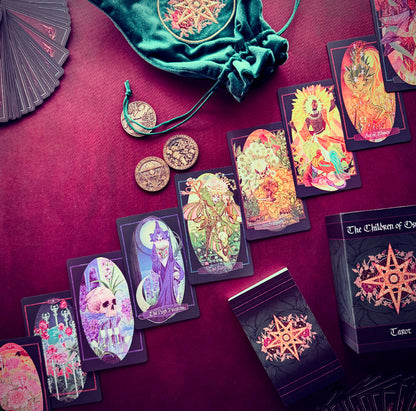A styled flat-lay of a tarot deck set on a deep burgundy fabric background, featuring illustrated tarot cards, a green velvet drawstring bag, several gold-toned coins, and the tarot box. The cards display colorful fantasy and gothic illustrations, and the box is labeled 'The Children of Osiris Tarot'.