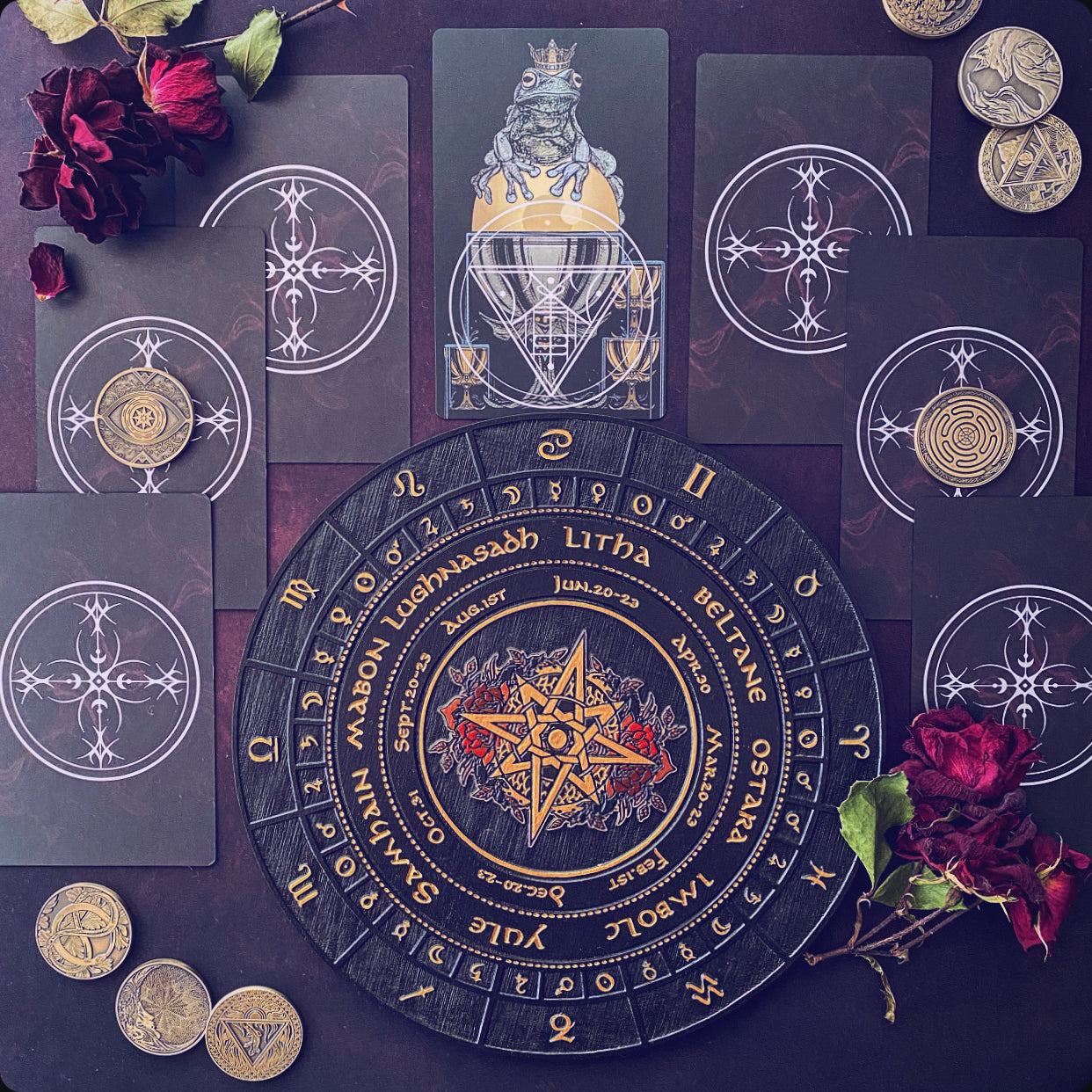 A flat-lay arrangement of occult-themed tarot cards and a circular divination board on a dark background, accented with red roses and gold coins. The central board features a gold star and the words 'Litha Septembris' and 'Januarius,' surrounded by dark cards with white mystical symbols.