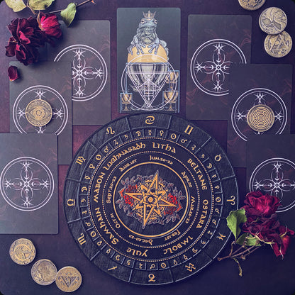 A flat-lay arrangement of occult-themed tarot cards and a circular divination board on a dark background, accented with red roses and gold coins. The central board features a gold star and the words 'Litha Septembris' and 'Januarius,' surrounded by dark cards with white mystical symbols.