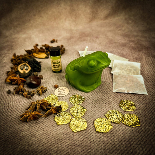 Green frog candle, bottle, gold coins, and dried herbs on a textured surface