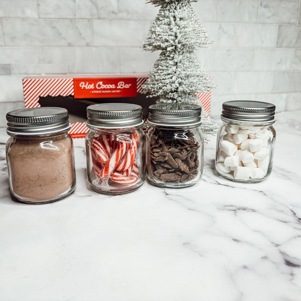 Four jars with hot cocoa ingredients on a marble surface with a small Christmas tree in the background.