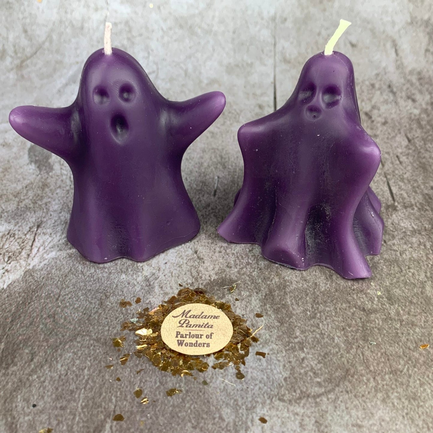 Two purple ghost-shaped candles on a gray surface with a gold label.