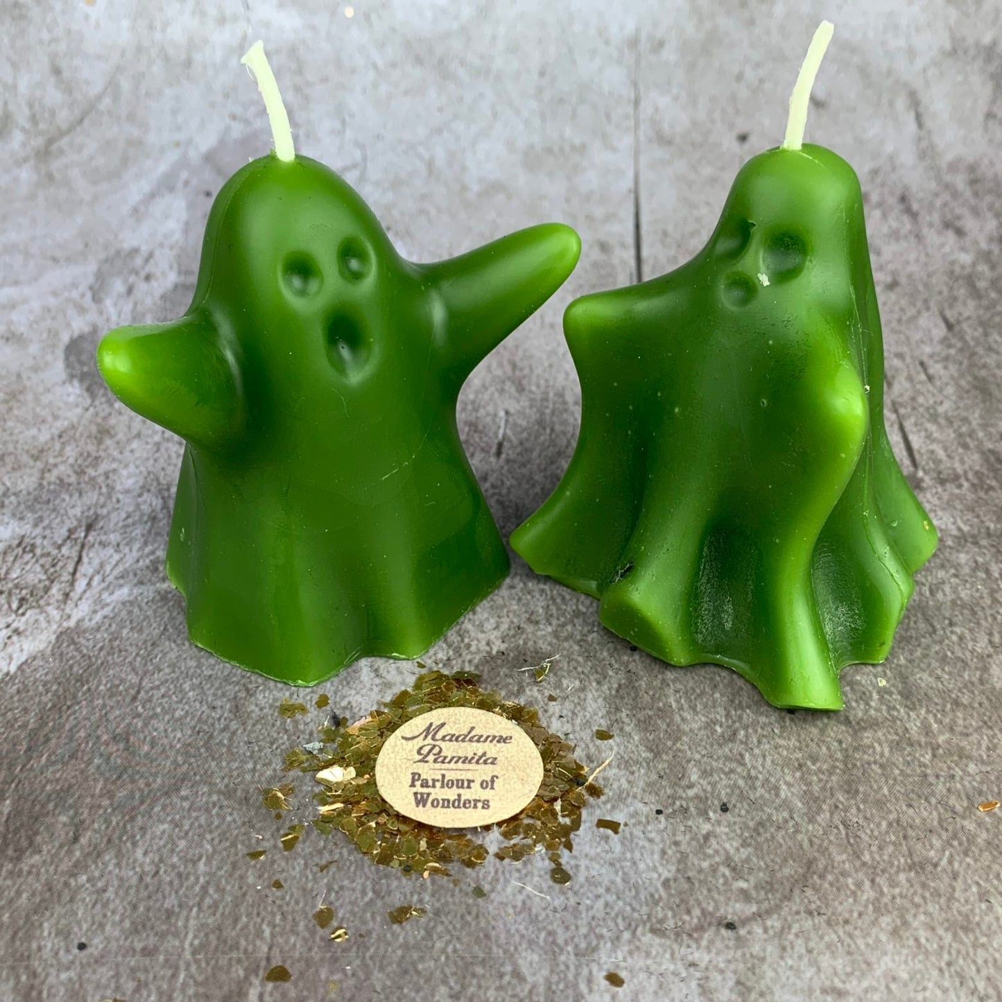Two green ghost-shaped candles on a gray surface with a gold label.