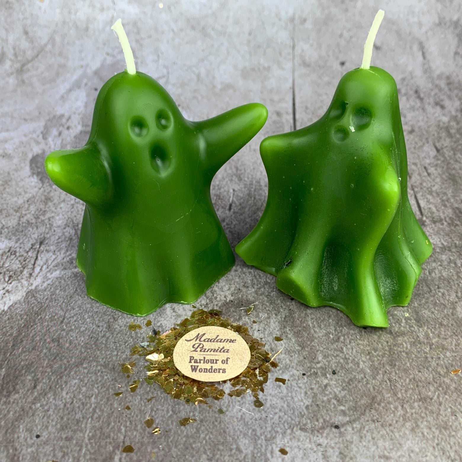 Two green ghost-shaped candles on a gray surface with a gold label.