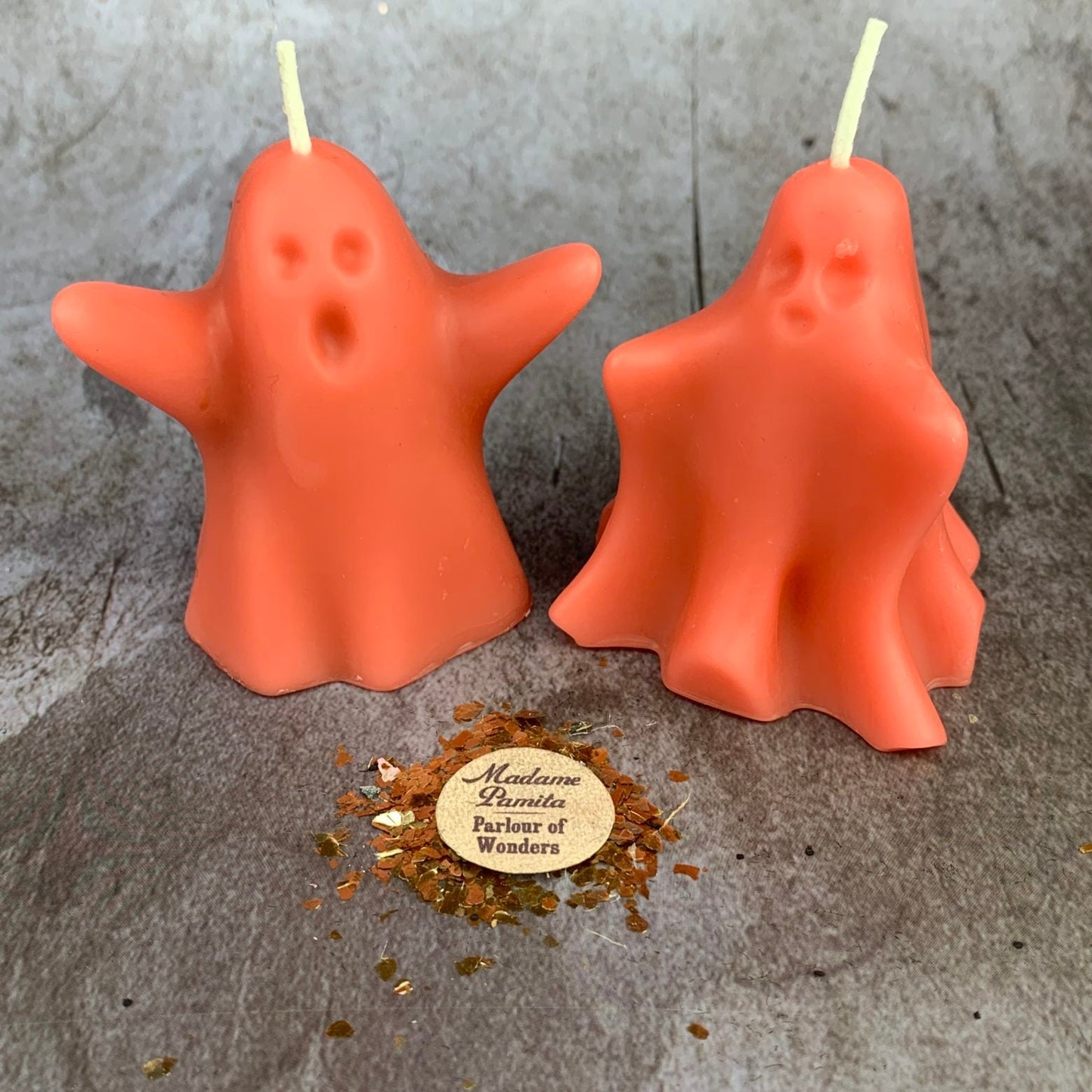 Two ghost-shaped candles on a textured surface with a brand label in the foreground.