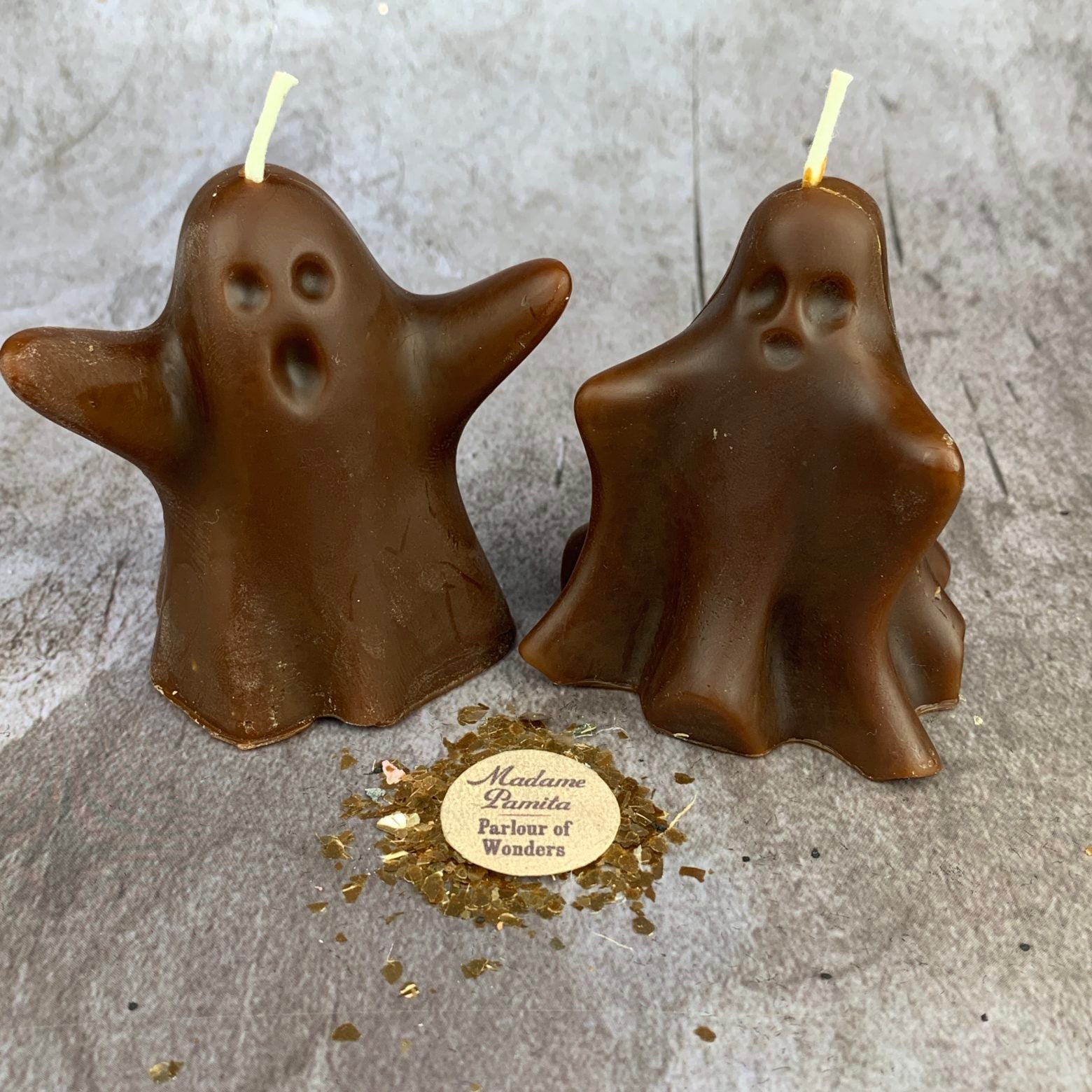 Two brown ghost-shaped candles on a gray surface with a gold label underneath.