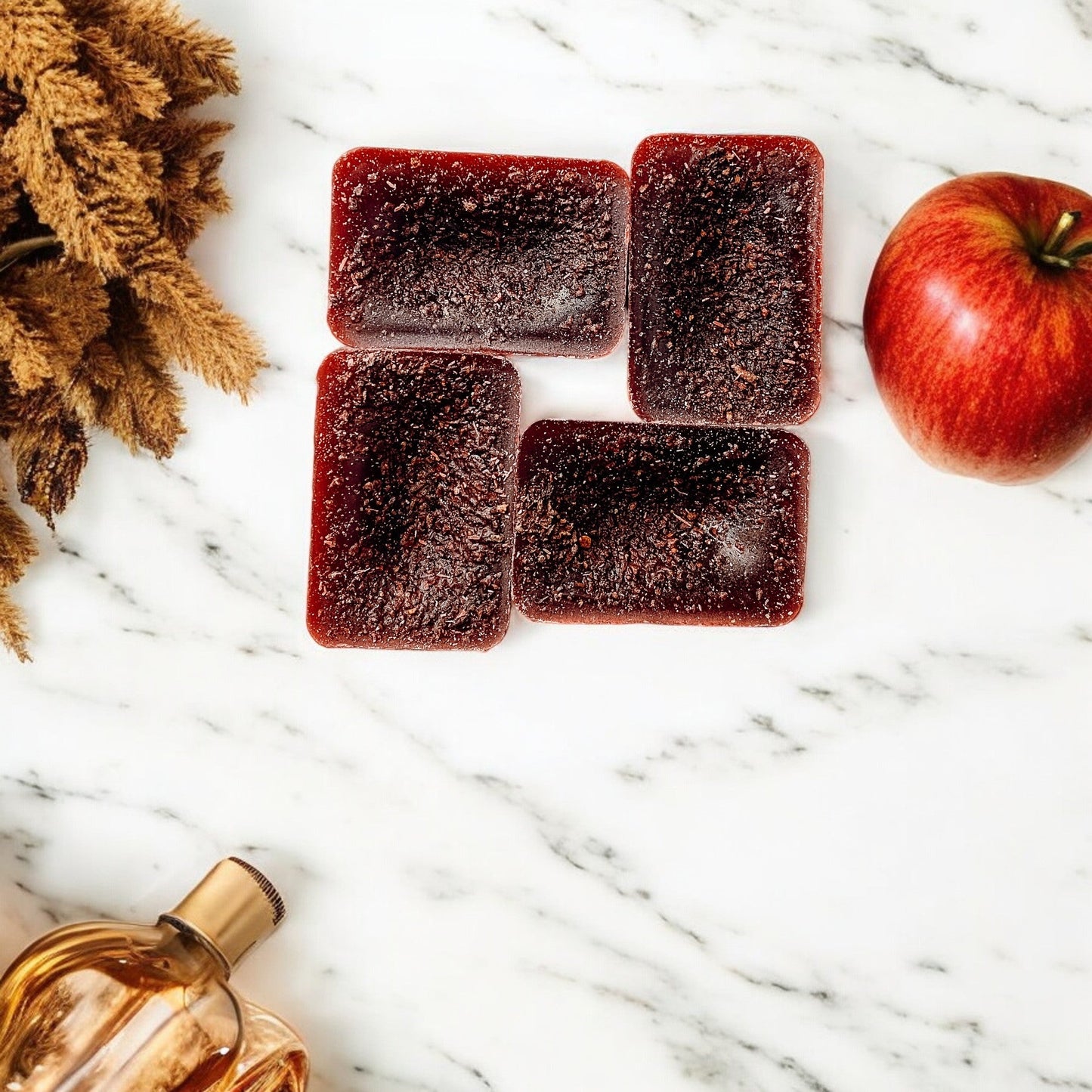 Five square red fruit gummies on a marble surface with an apple and perfume bottle.