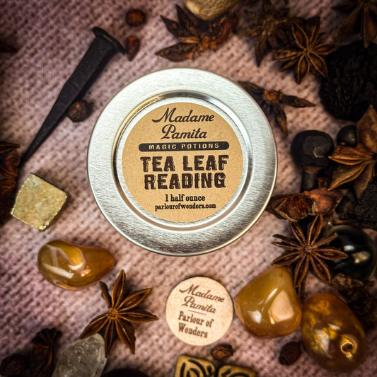 Metallic tin labeled 'Tea Leaf Reading' surrounded by dried herbs and stones on a textured surface.