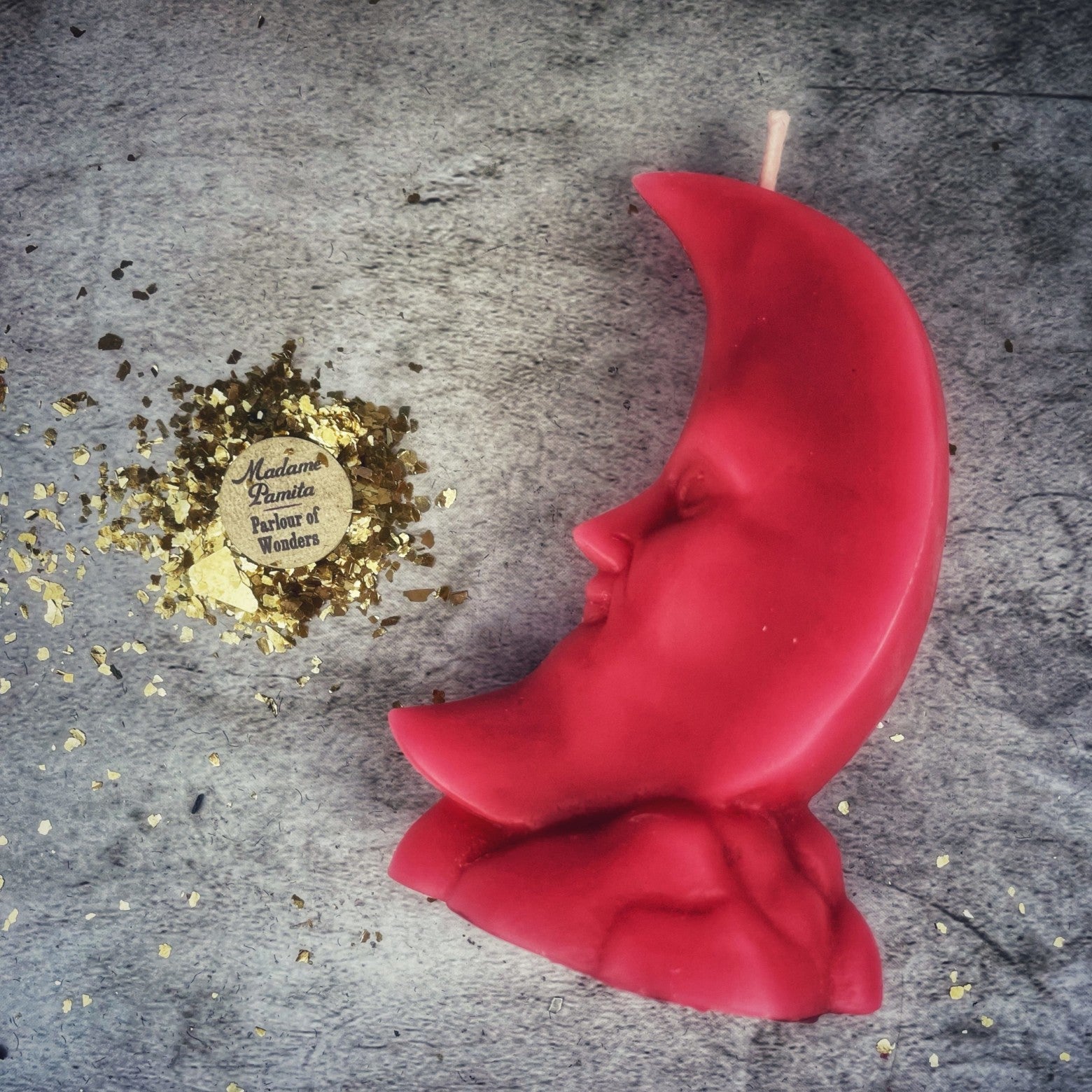 Red moon-shaped candle on a textured surface with gold confetti and a logo.