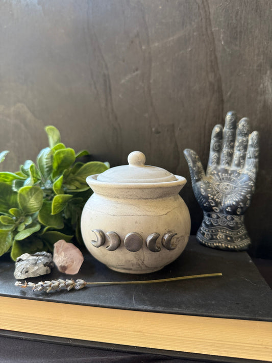 Decorative stone jar with moon phase symbols on a dark surface with plants and a palm reading hand.