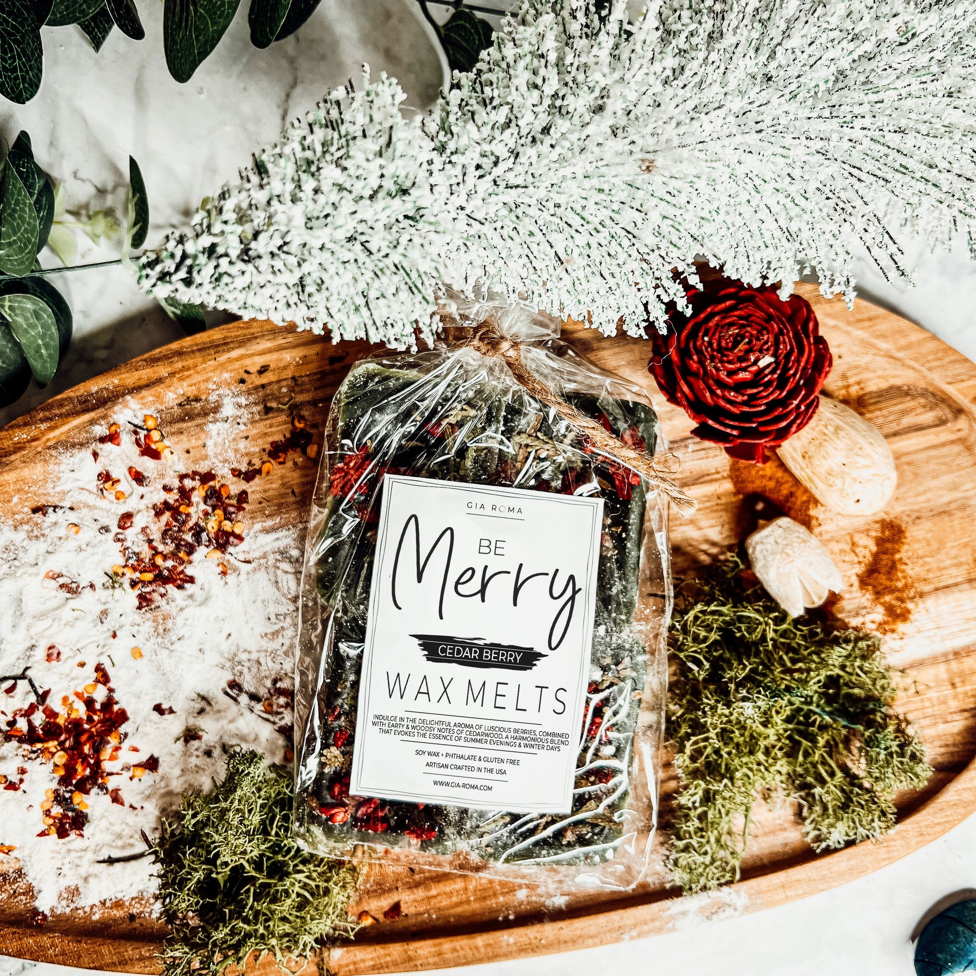 Packaged wax melts labeled 'Be Merry' on a decorative wooden surface with greenery and flowers.