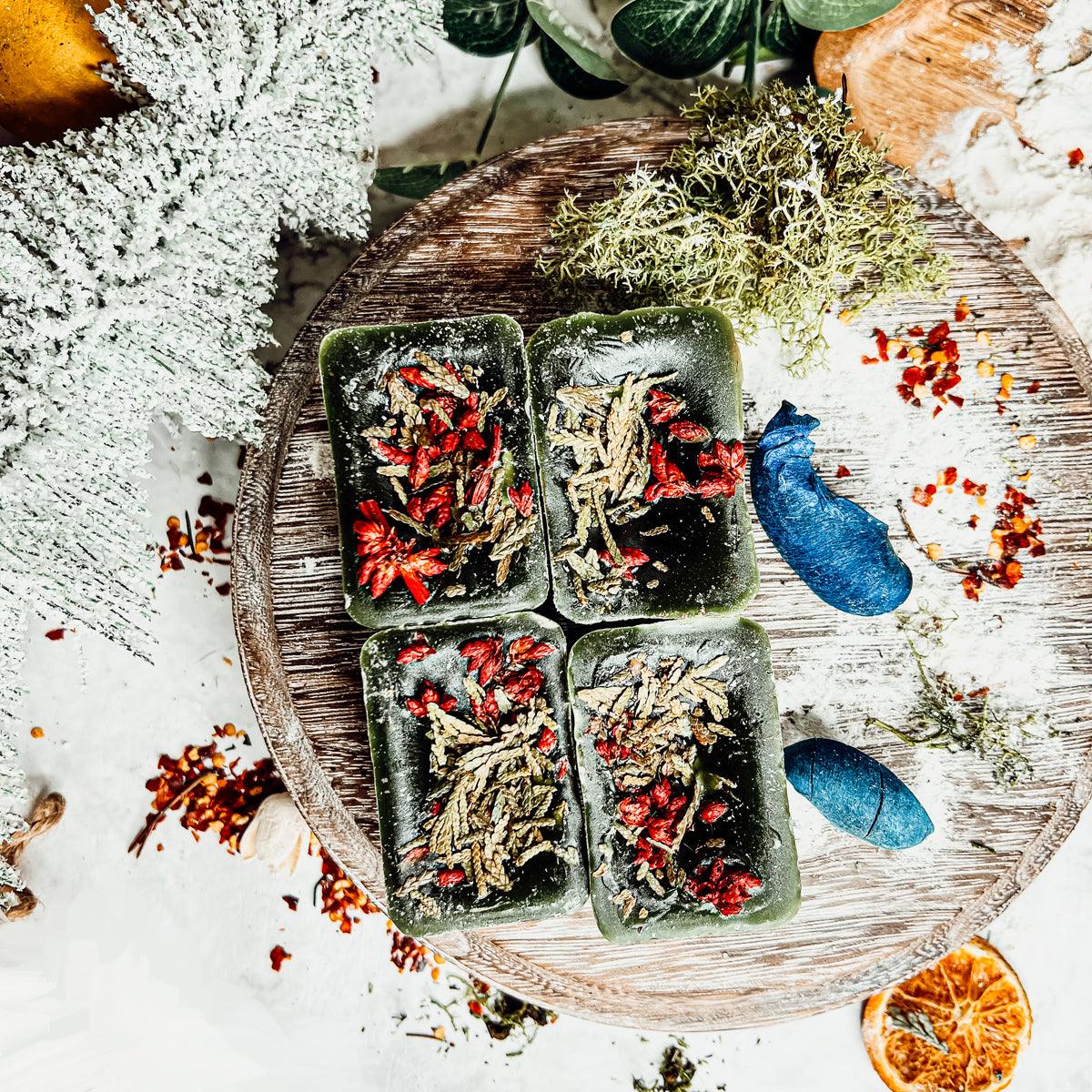 Four rectangular green soap bars with dried herbs on a wooden board with decorative elements.