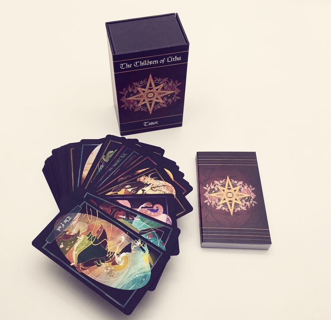 A deck of illustrated oracle cards with a matching box, shown on a plain light background. The box and card backs are dark purple with a gold star design, while the fanned cards reveal colorful fantasy-style artwork.