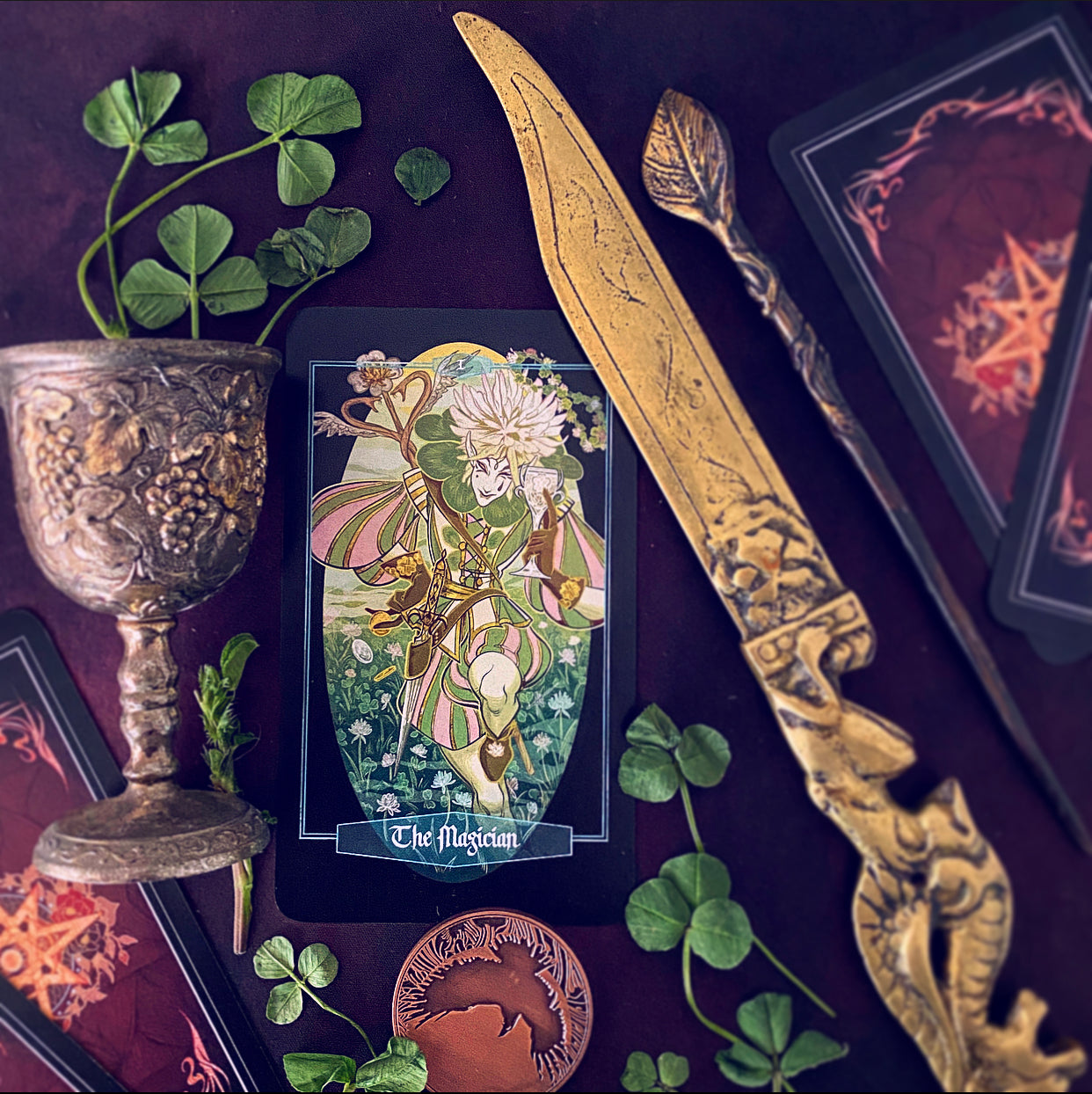 A styled flat-lay featuring a central illustrated tarot card labeled 'The Magician,' surrounded by decorative props including a large ornate gold knife, a silver goblet with green clover, a bronze coin, and dark red tarot card backs on a deep purple background.