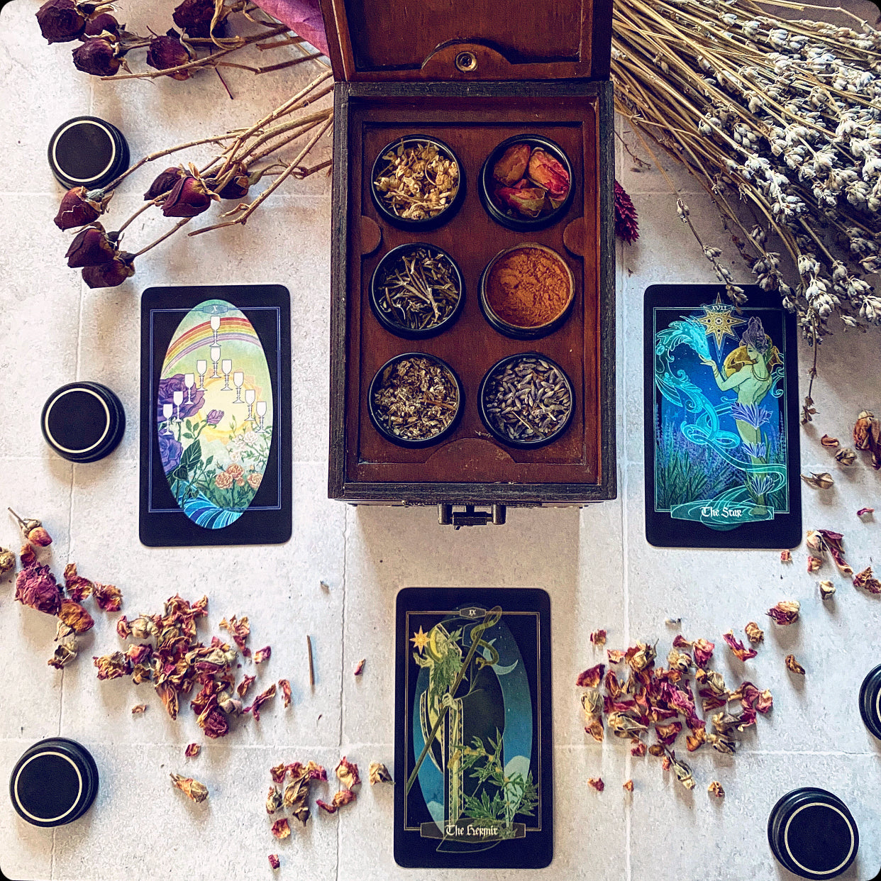An open wooden box filled with small bowls of dried botanicals is arranged on a light surface with three illustrated tarot cards, loose dried flowers, and small black cups in a styled flat-lay composition.