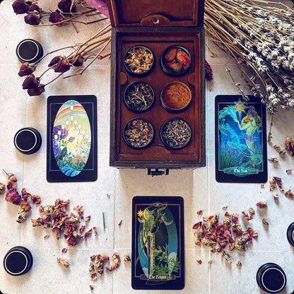 An open wooden box filled with small bowls of dried botanicals is arranged on a light surface with three illustrated tarot cards, loose dried flowers, and small black cups in a styled flat-lay composition.