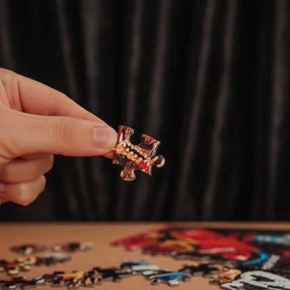 A close-up of a hand holding a small, intricately designed jigsaw puzzle piece with a red, black, and gold pattern, with a blurred jigsaw puzzle box and loose pieces in the background.