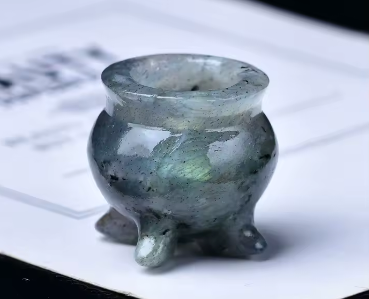 A small polished stone censer or tripod vessel with a rounded body and three short feet, shown close up on a white printed surface. The material appears translucent smoky gray-green with subtle iridescent flecks.