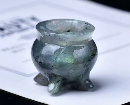 A small polished stone censer or tripod vessel with a rounded body and three short feet, shown close up on a white printed surface. The material appears translucent smoky gray-green with subtle iridescent flecks.