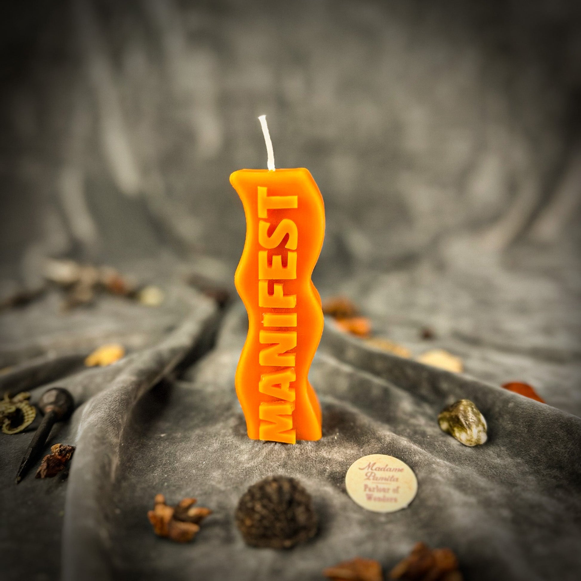 Orange candle shaped like a gun with 'MANIFEST' text on a textured surface with decorative elements.