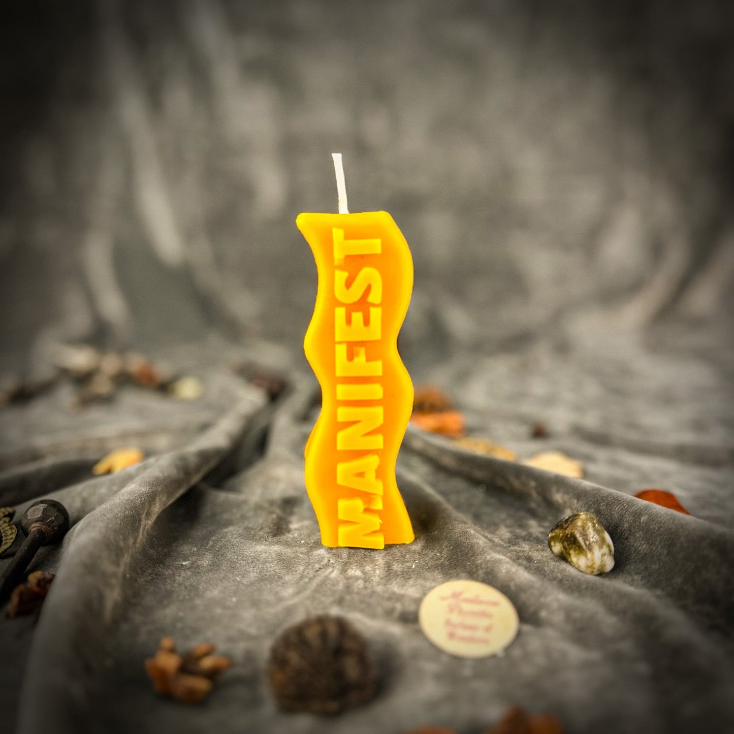 Yellow candle with 'MANIFEST' text on a textured surface with stones and coins.