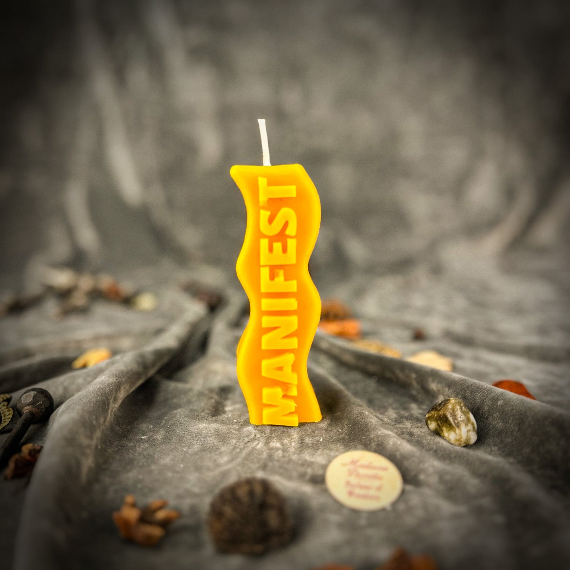 Yellow candle with 'MANIFEST' text on a textured surface with stones and coins.