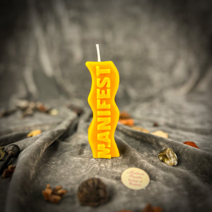 Yellow candle with 'MANIFEST' text on a textured surface with stones and coins.