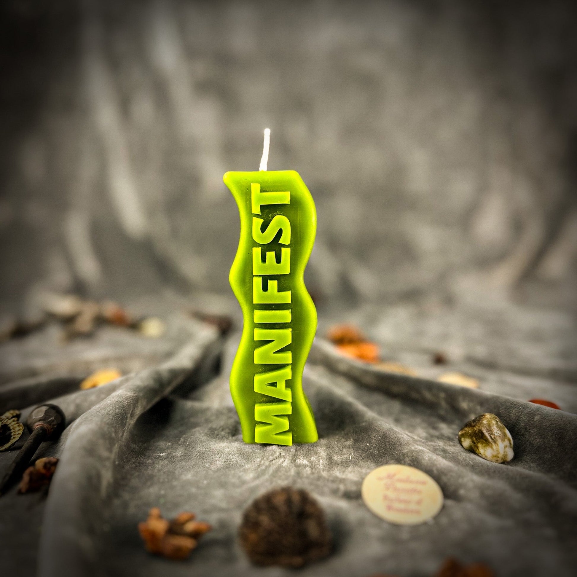 Green candle with 'MANIFEST' text on a dark fabric background